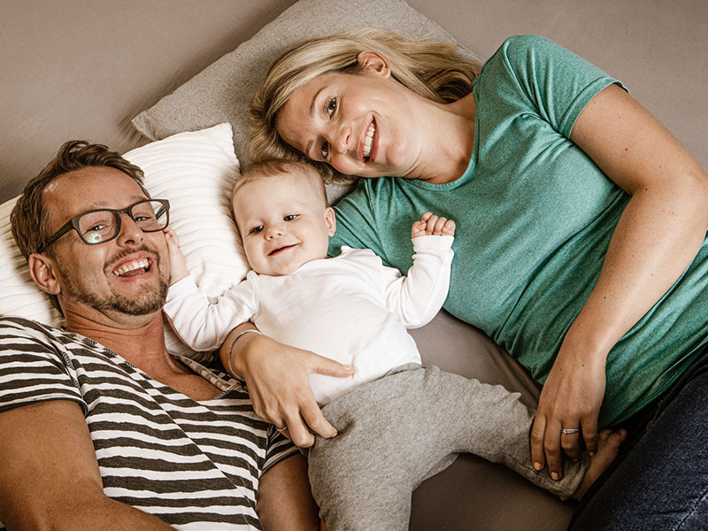 Eine Familie mit Kleinkind liegt zusammen im Bett und lächelt in die Kamera